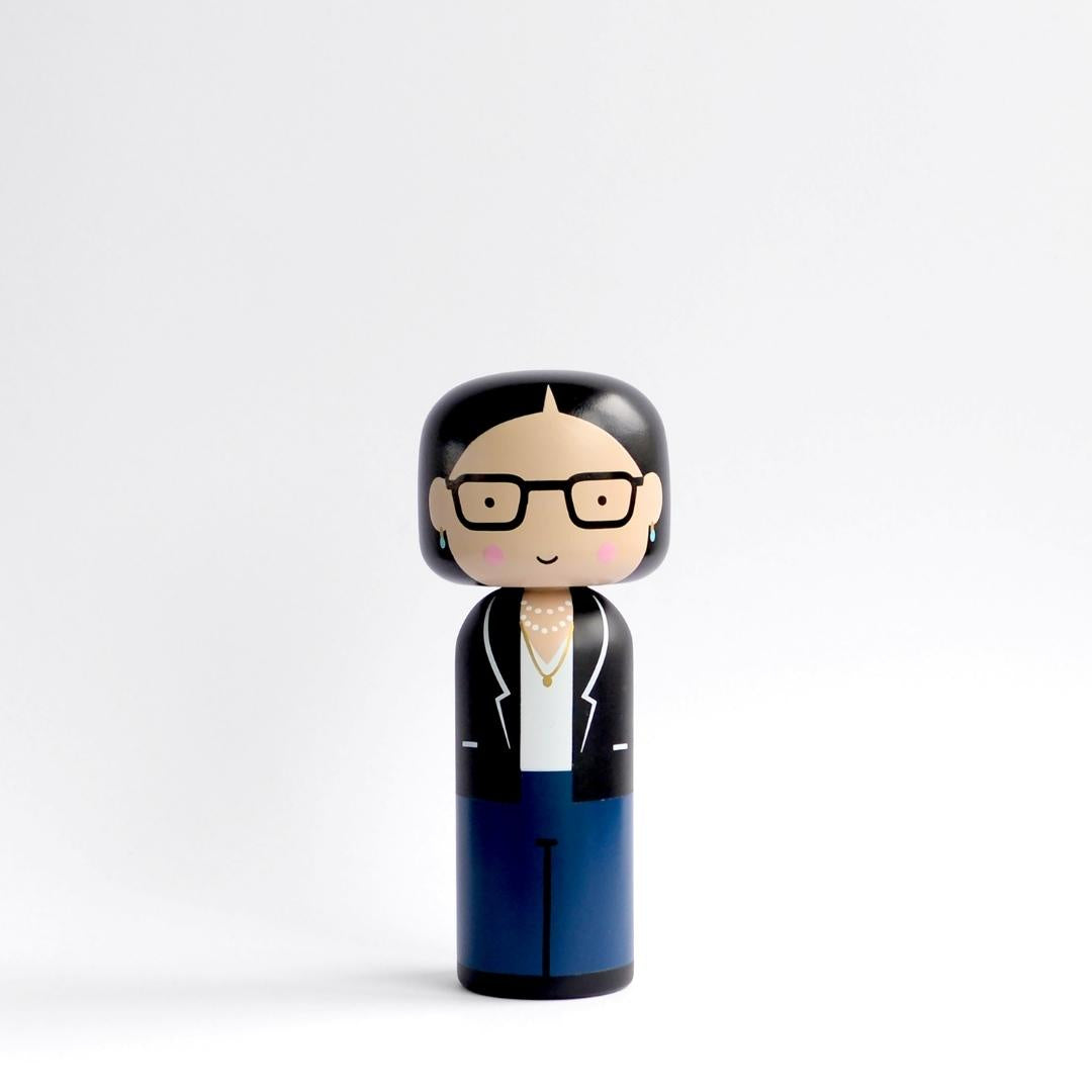 En kokeshi-dukke med briller, sort hår, sort blazer, hvid skjorte og blå bukser på en hvid baggrund.