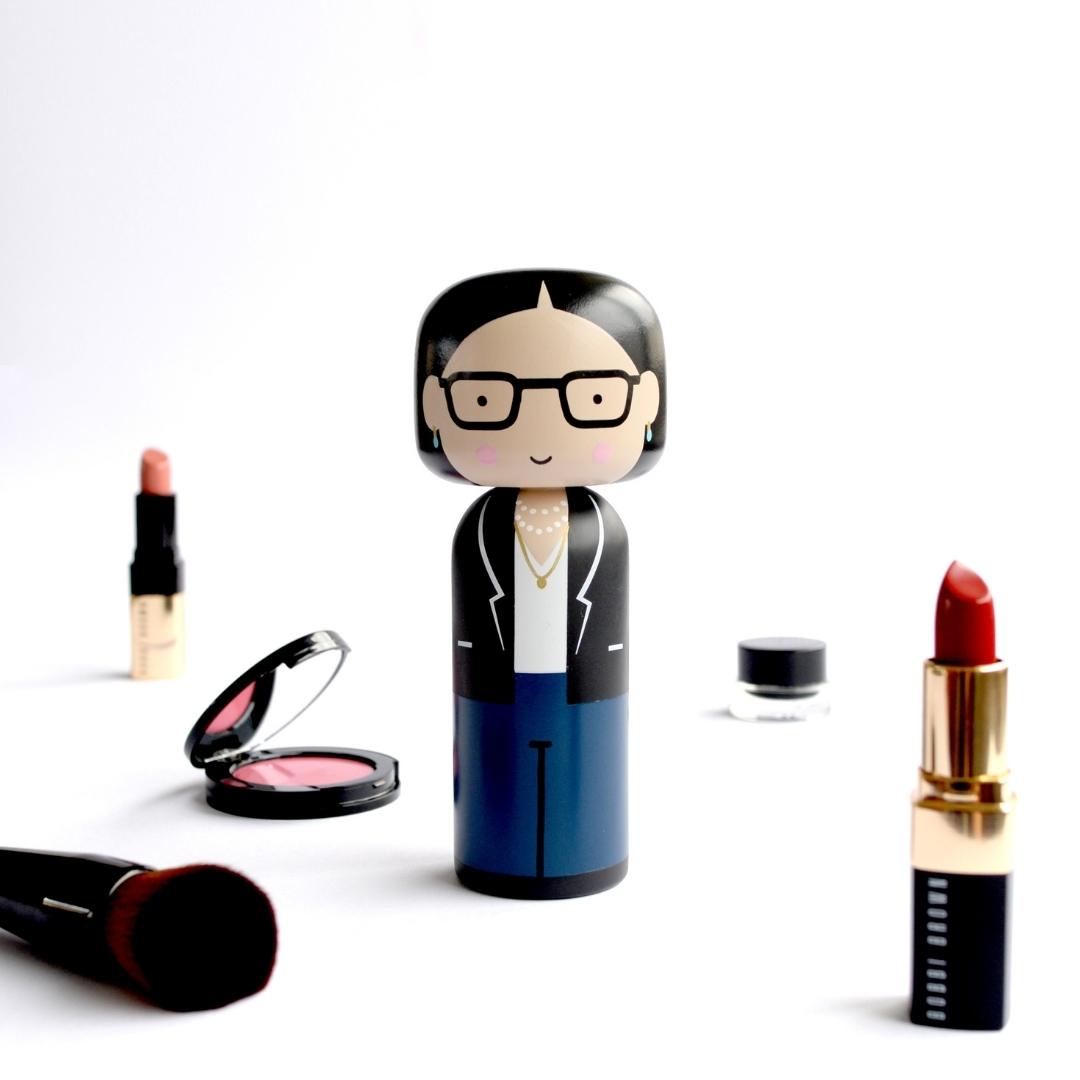 En kokeshi-dukke med briller og jakkesæt, omgivet af makeup-artikler på en hvid baggrund.