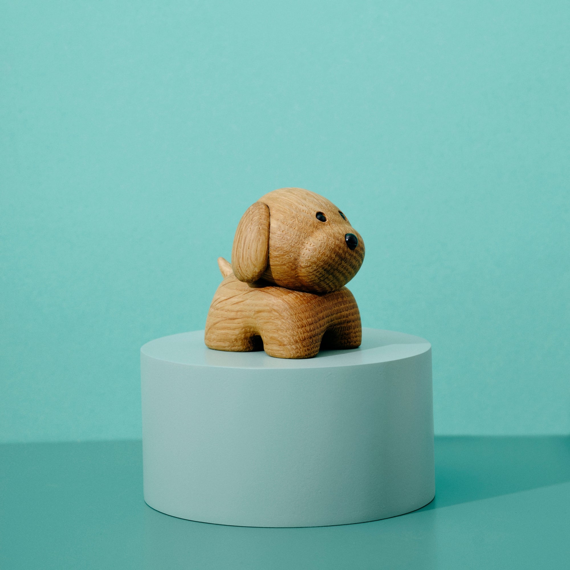 En lille Figurer af træ på en lyseblå cylindrisk sokkel mod en blågrøn baggrund.