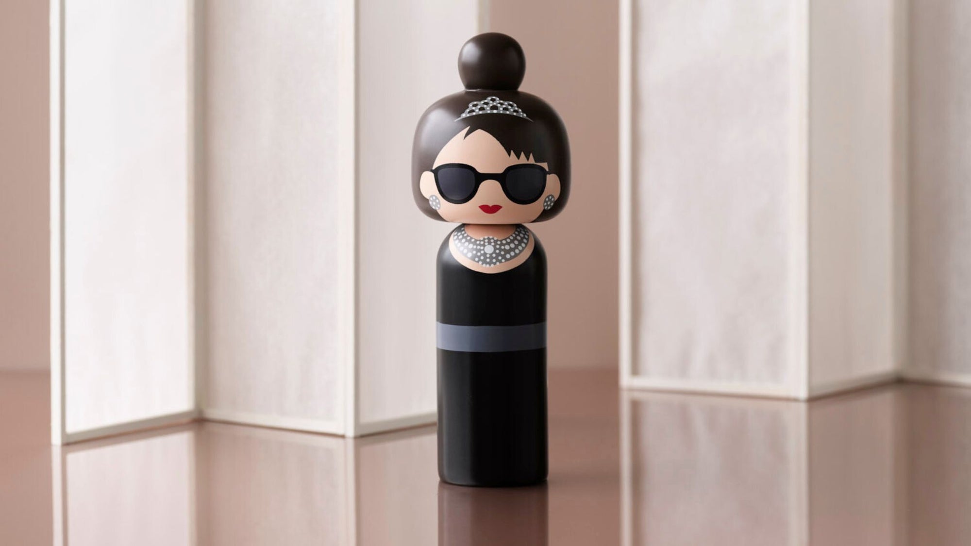 En kokeshi-dukke med solbriller, tiara og perlekæde på en reflekterende overflade.