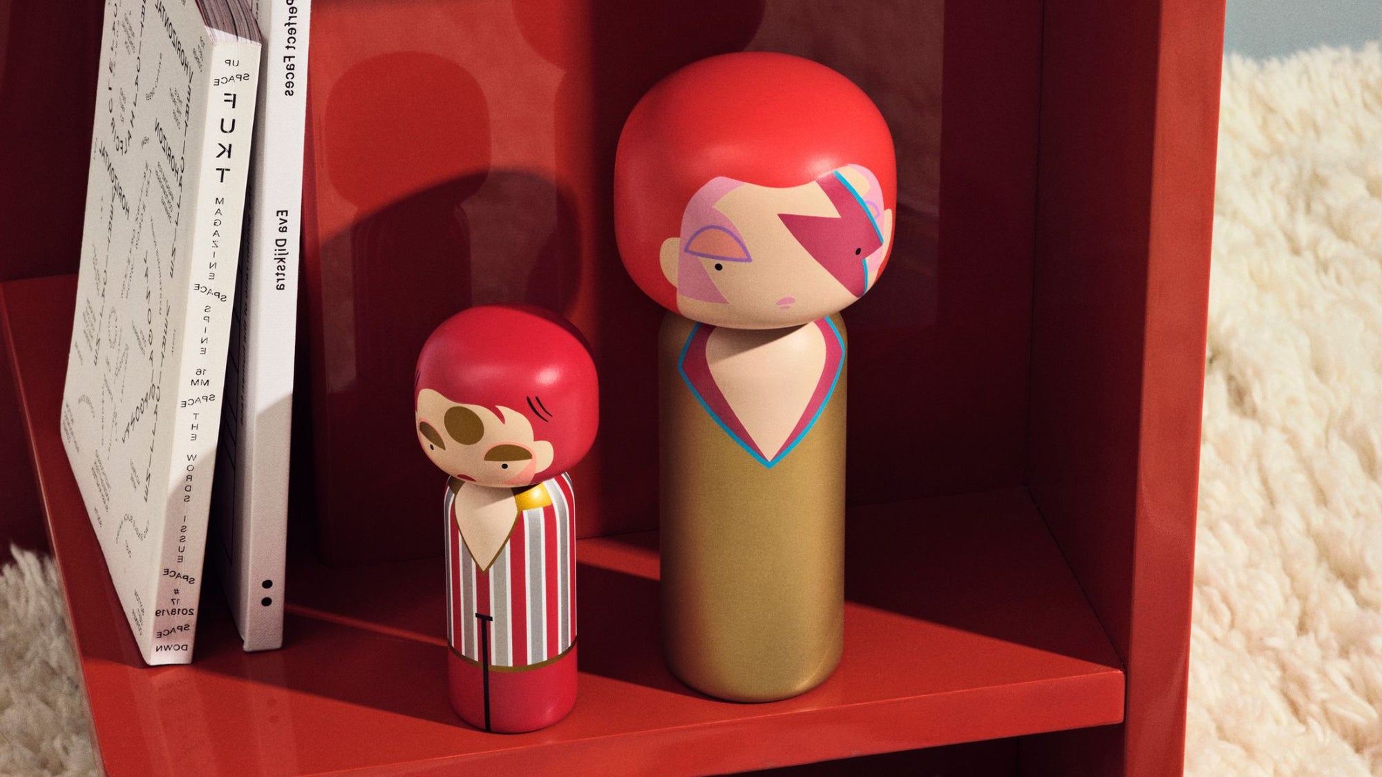 To farverige Kokeshi Dukker på en rød hylde ved siden af en stak bøger.