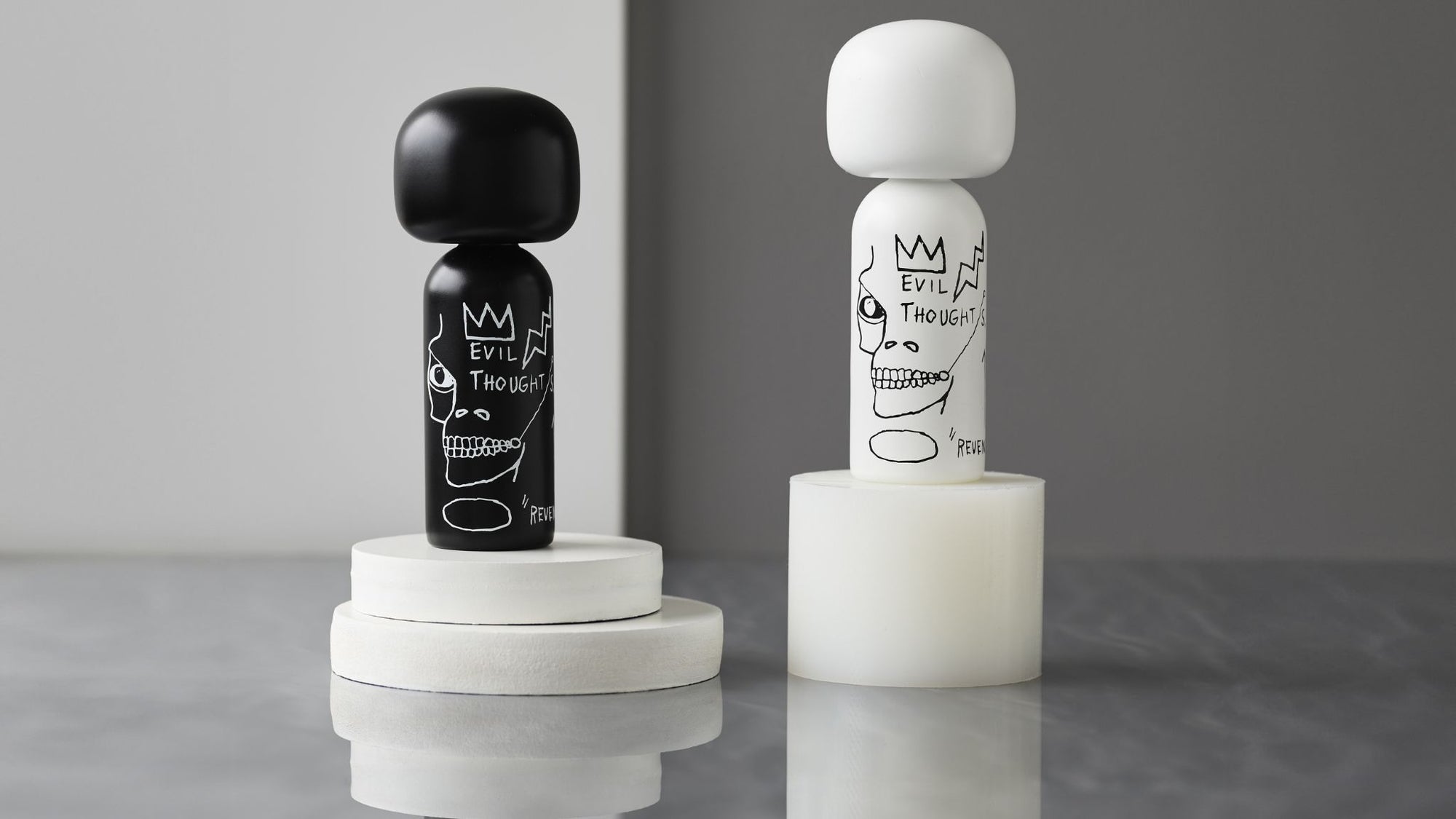 To Kokeshi Dukker, en sort og en hvid, med kunstneriske motiver, på runde sokler mod en grå baggrund.
