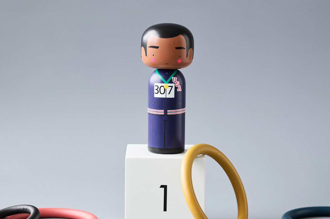 Kokeshi-dukke i blåt tøj med "307" på en hvid sokkel mærket "1", omgivet af farverige ringe.