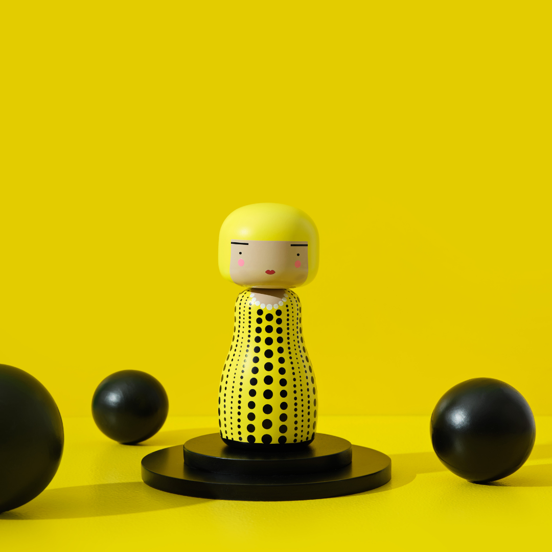 Kokeshi-dukke med gult hår og dot kjole på et sort stativ, omgivet af sorte kugler.