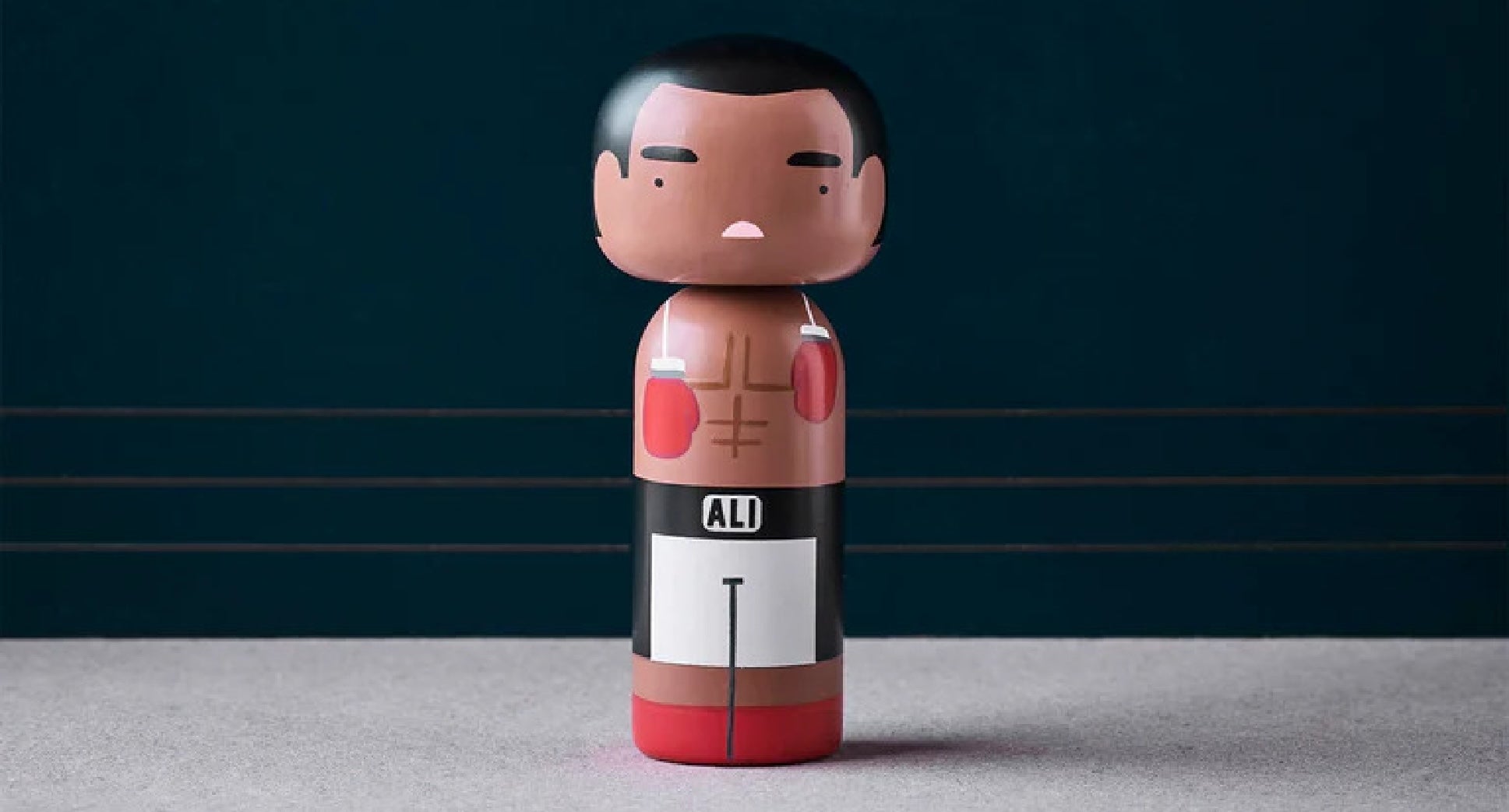 En kokeshi-dukke, der ligner en bokser med røde handsker og hvide shorts, står på en grå overflade.