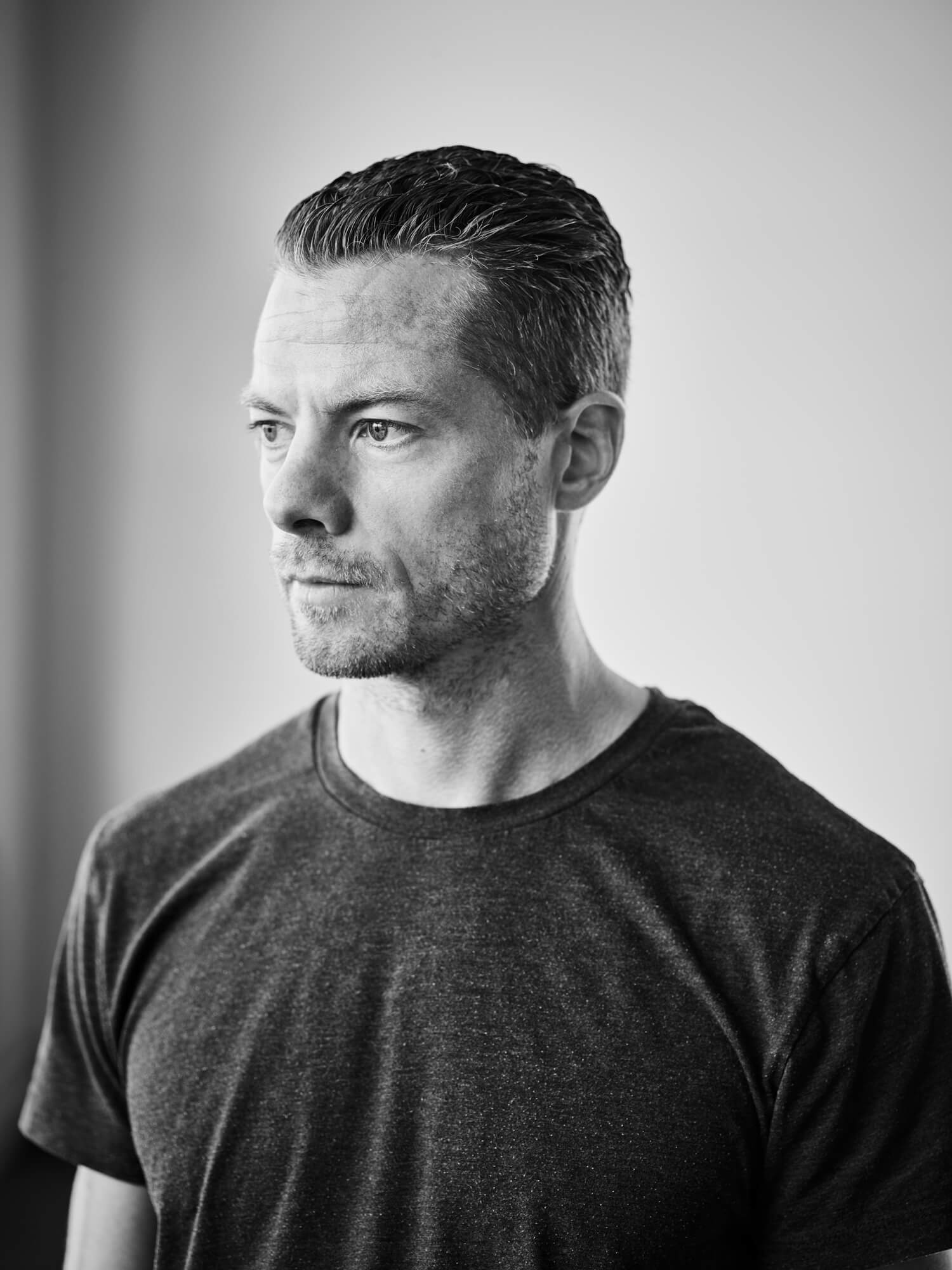 En person med kort hår og skæg, iført en mørk t-shirt, kigger til siden på et sort-hvidt foto.