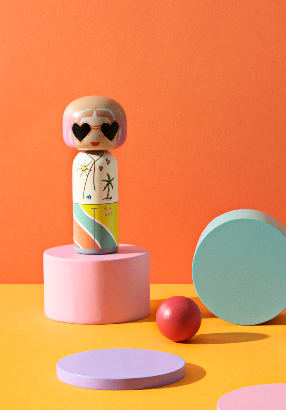 Kokeshi-dukke med hjerteformede solbriller på farverige geometriske figurer.