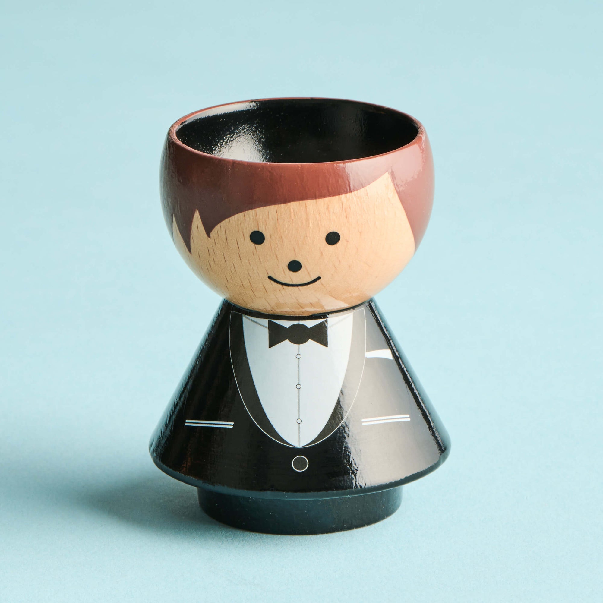 Eine hölzerne Kokeshi-Puppe mit Smoking-Design und einem lächelnden Gesicht auf hellblauem Hintergrund.