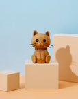 Eine hölzerne Katzenfigur mit schwarzen Schnurrhaaren sitzt auf einem beigen Block vor einem pastellblauen und pfirsichfarbenen Hintergrund.