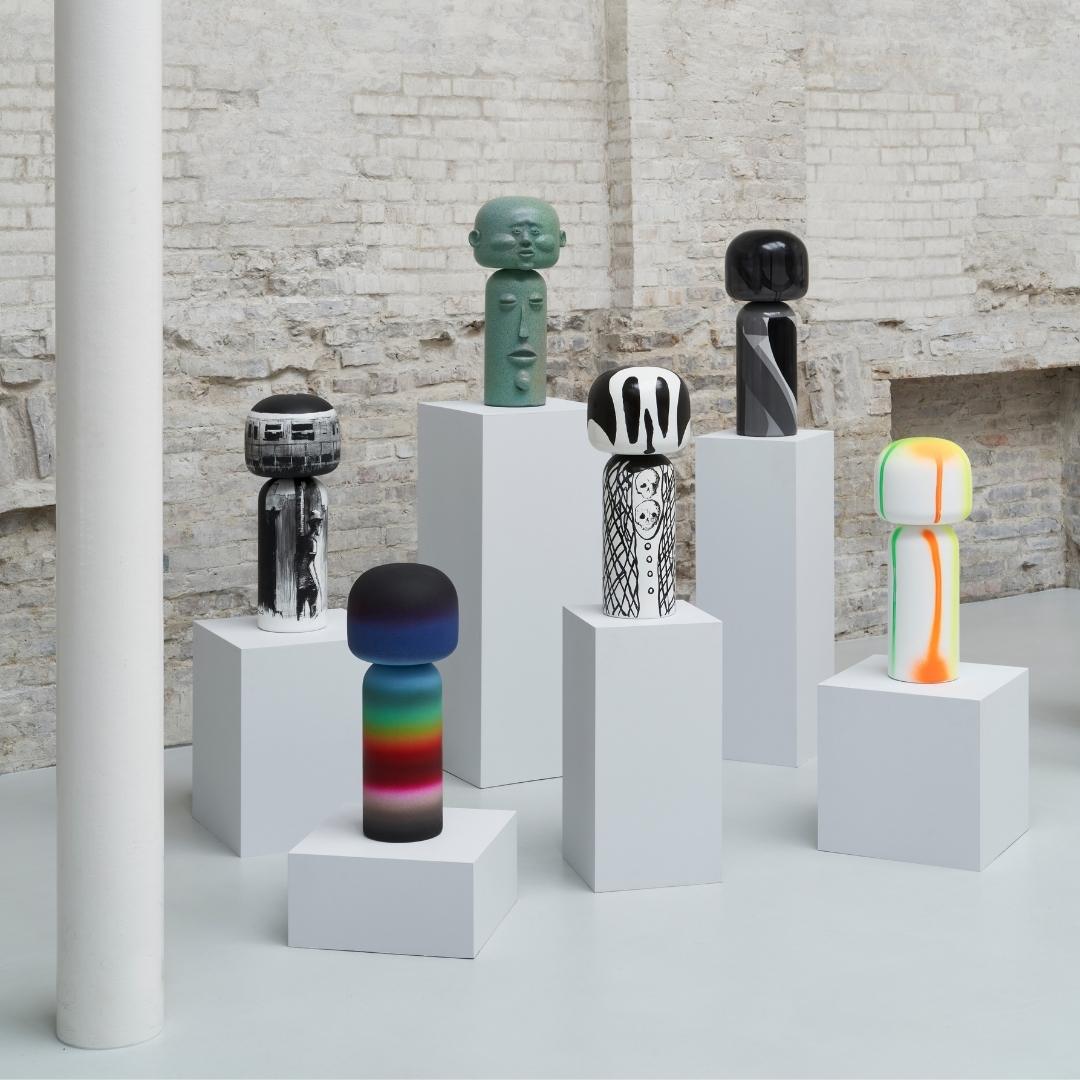 Kokeshi-Puppen mit verschiedenen Motiven auf weißen Sockeln in einem minimalistischen Raum mit Backsteinwänden.