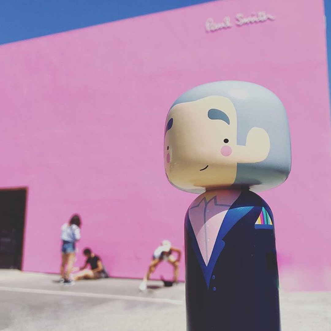 Eine Kokeshi-Puppe in einem Anzug steht vor einer leuchtend rosa Wand mit Menschen im Hintergrund.