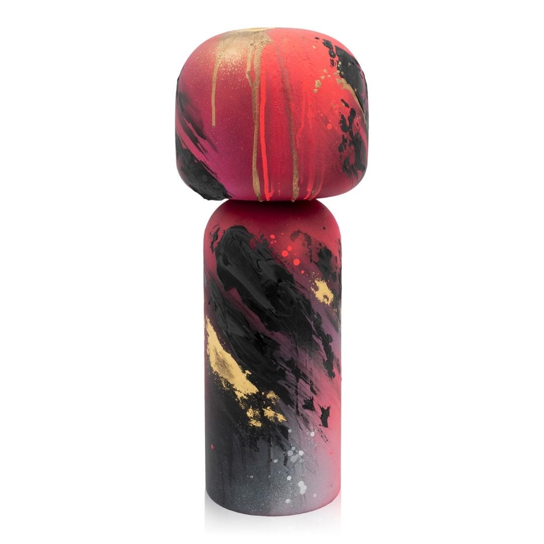 Eine bunte Kokeshi-Puppe mit abstrakten roten, schwarzen und goldenen Farbspritzern auf einem zylindrischen Körper.