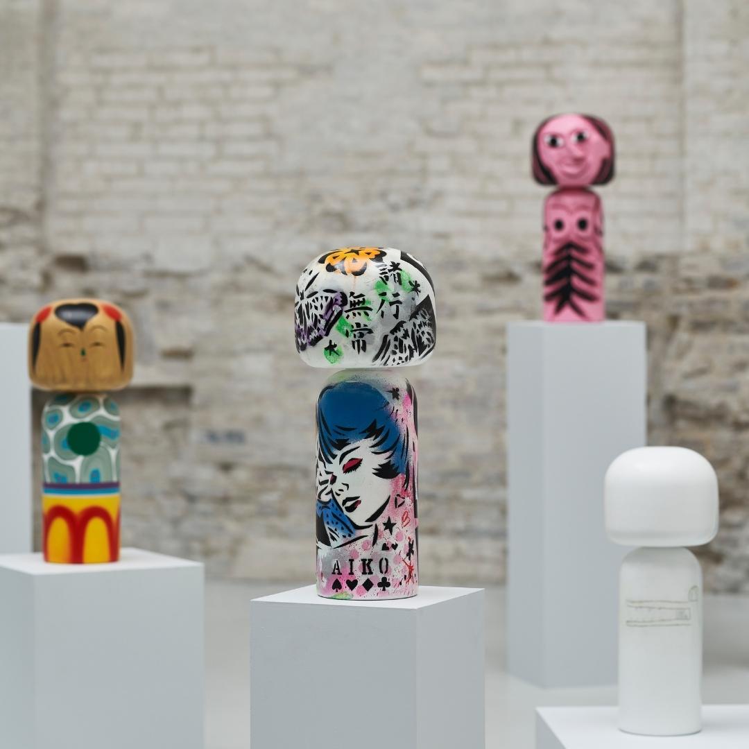 Kokeshi-Puppen mit verschiedenen Motiven auf weißen Sockeln vor einem Backsteinmauer-Hintergrund.