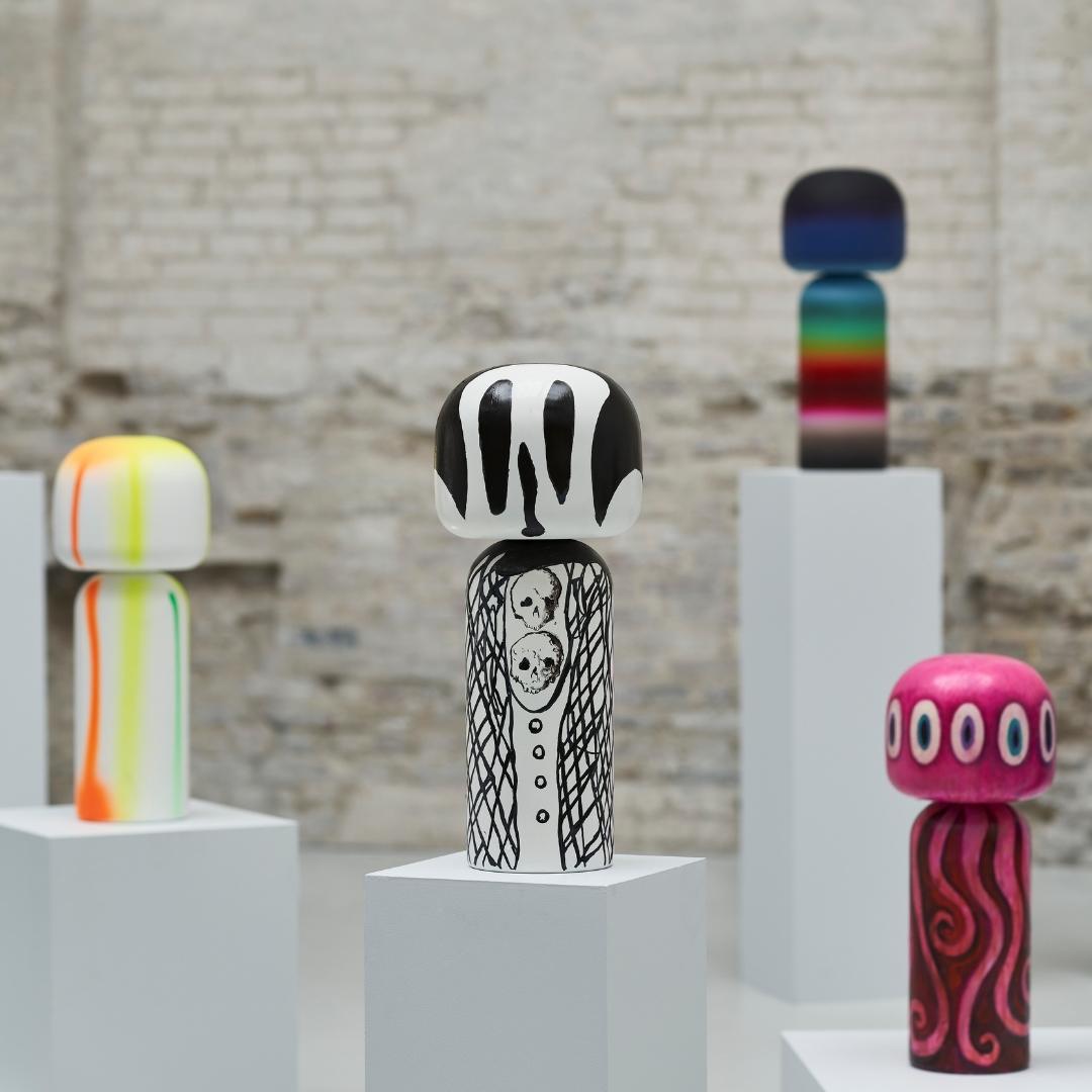 Vier bunte Kokeshi-Puppen auf Sockeln vor einem Backsteinmauer-Hintergrund.