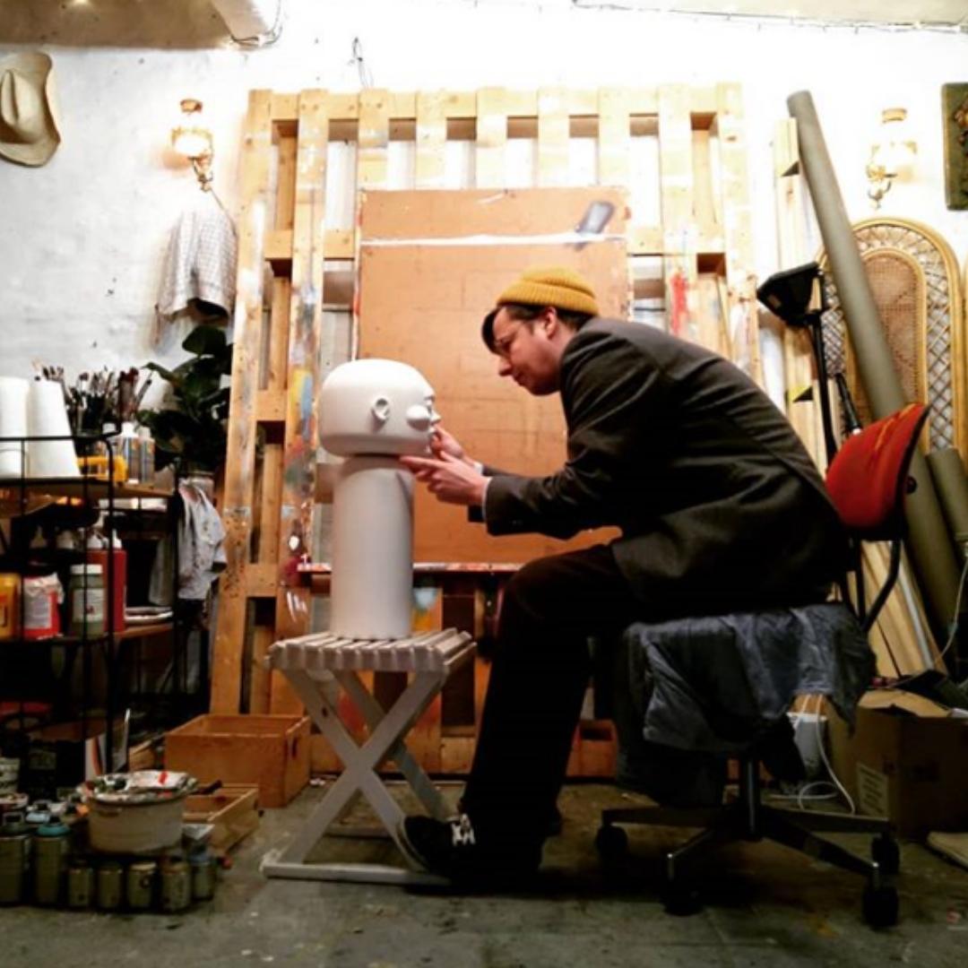 Eine Person in einer Werkstatt bemalt eine große Kokeshi-Puppe auf einem Hocker, umgeben von Kunstzubehör.