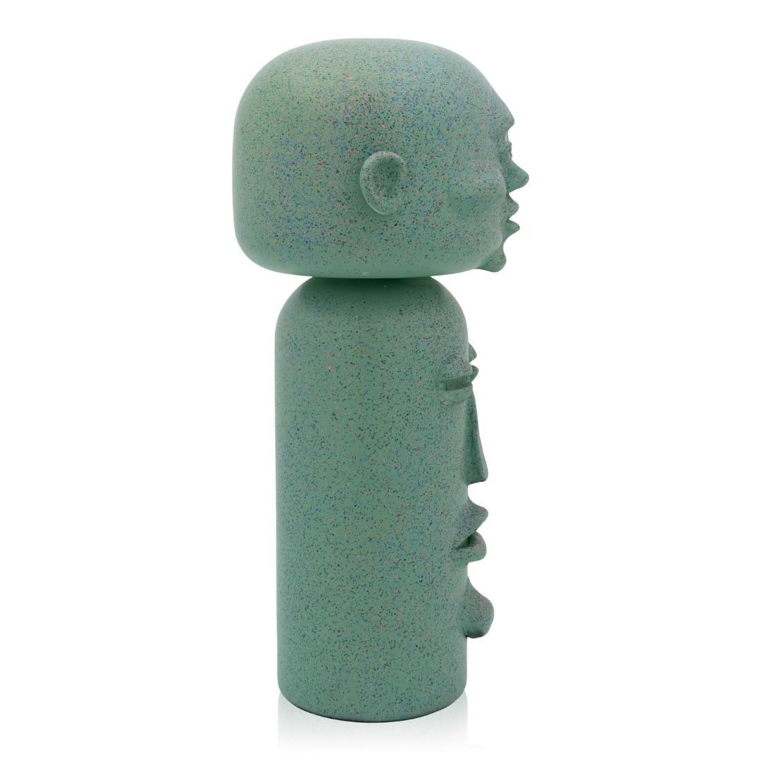 Eine grüne, gesprenkelte Kokeshi-Puppe mit abstrakten Gesichtszügen, von der Seite betrachtet.