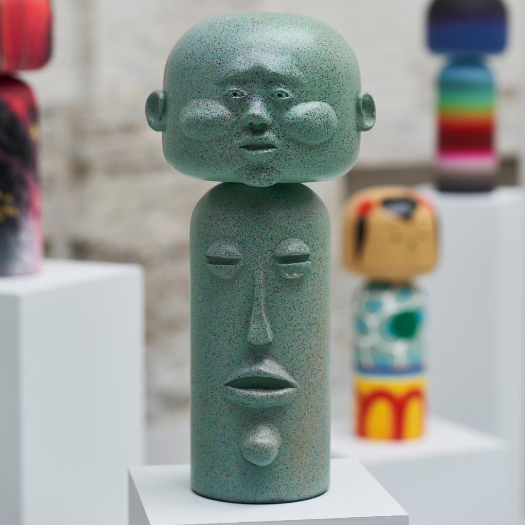 Eine grüne, abstrakte Kokeshi-Puppe mit einem großen Kopf und ausgestellten Gesichtszügen.