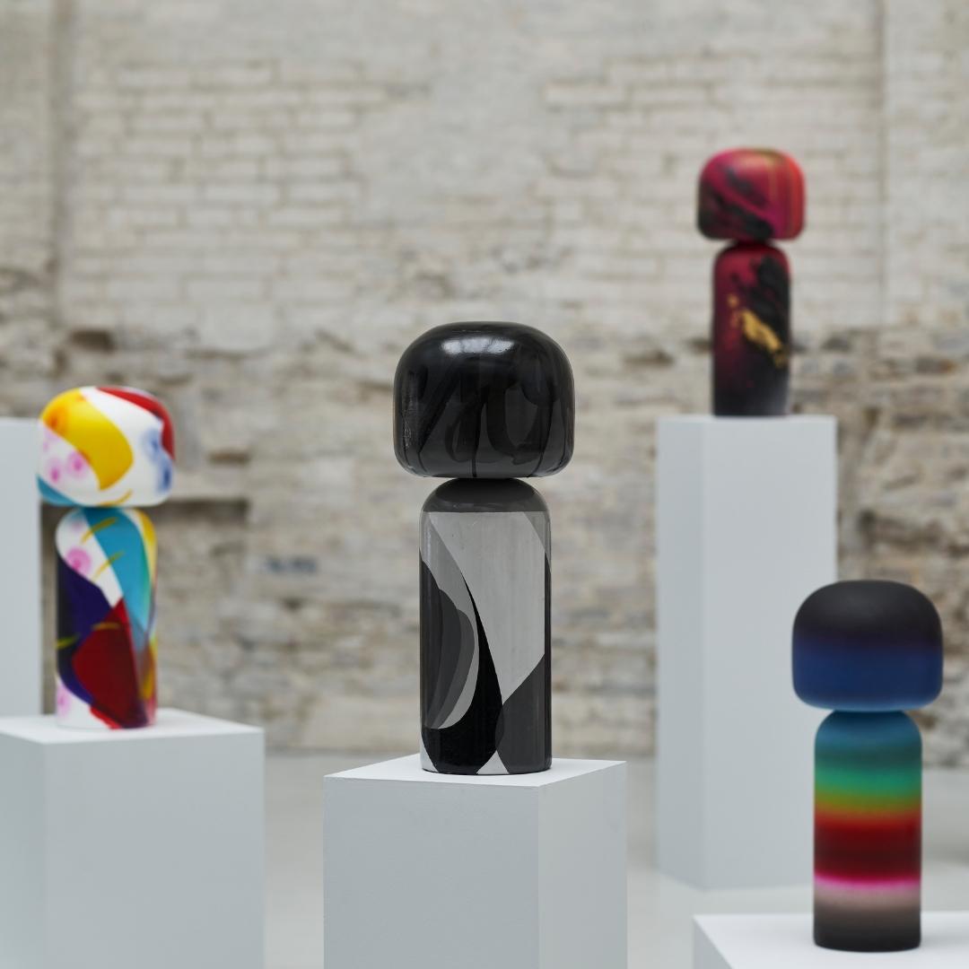 Bunte Kokeshi-Puppen auf weißen Sockeln vor einem Backsteinmauer-Hintergrund.
