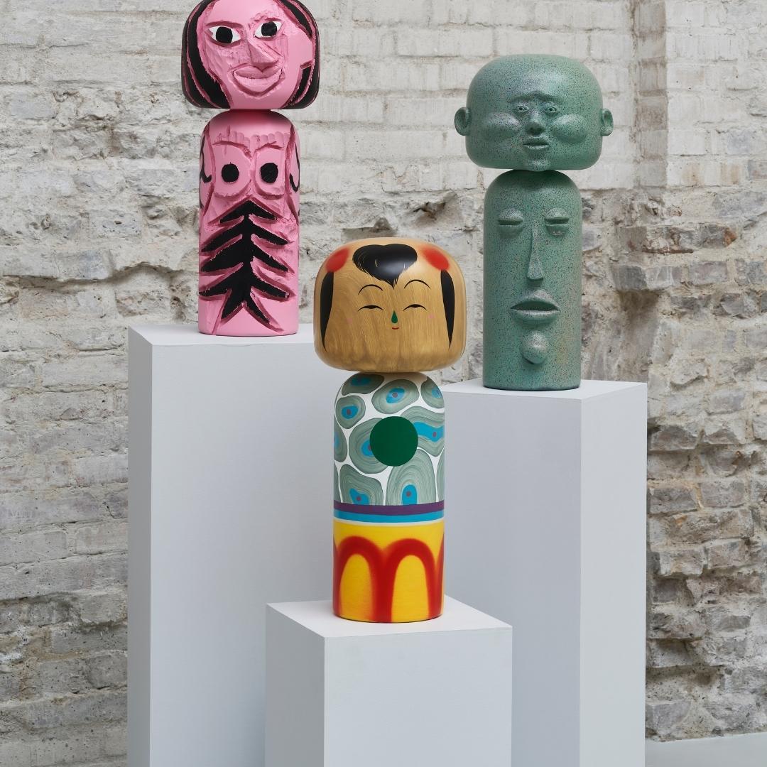 Drei bunte Kokeshi-Puppen auf weißen Sockeln vor einem strukturierten Backsteinmauer-Hintergrund.