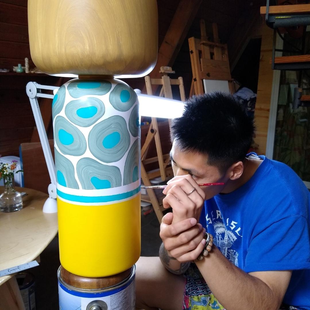 Künstlerin bemalt in einem Atelier eine große Kokeshi-Puppe mit einem blauen, grauen und gelben Muster.