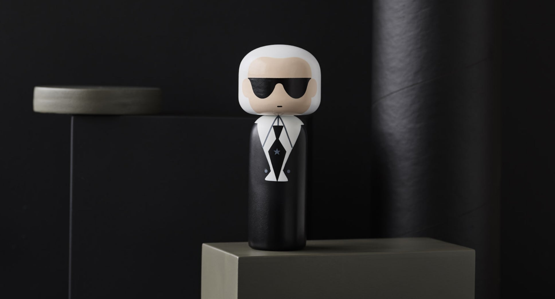 Eine Kokeshi-Puppe mit Sonnenbrille und Anzug steht auf einem Sockel vor einem dunklen Hintergrund.