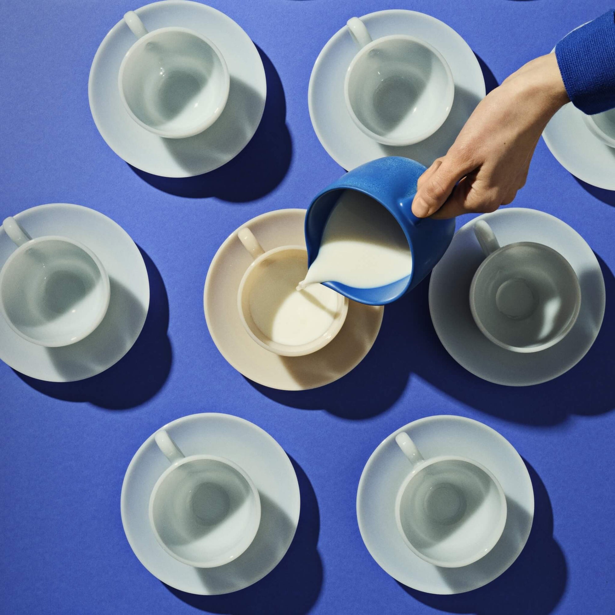 Eine Hand gießt milk aus einer blauen Kanne in eine Tasse, umgeben von leeren Tassen auf blauem Hintergrund.