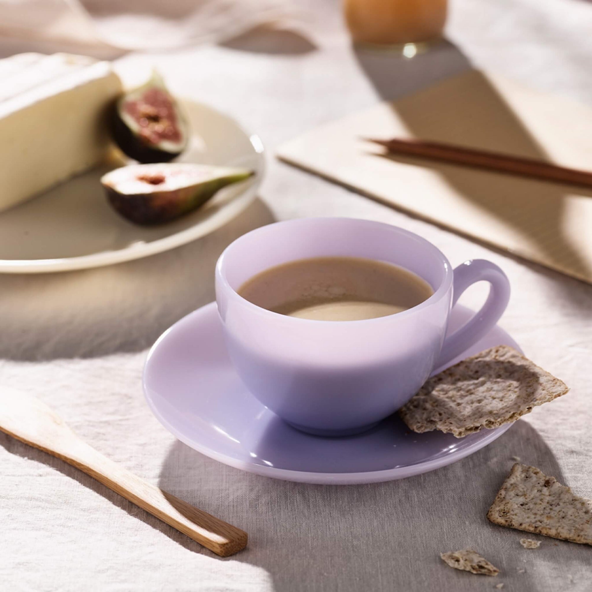 Eine Lavendeltasse Kaffee auf einer Untertasse mit Crackern, daneben ein Teller mit Käse und Feigen.