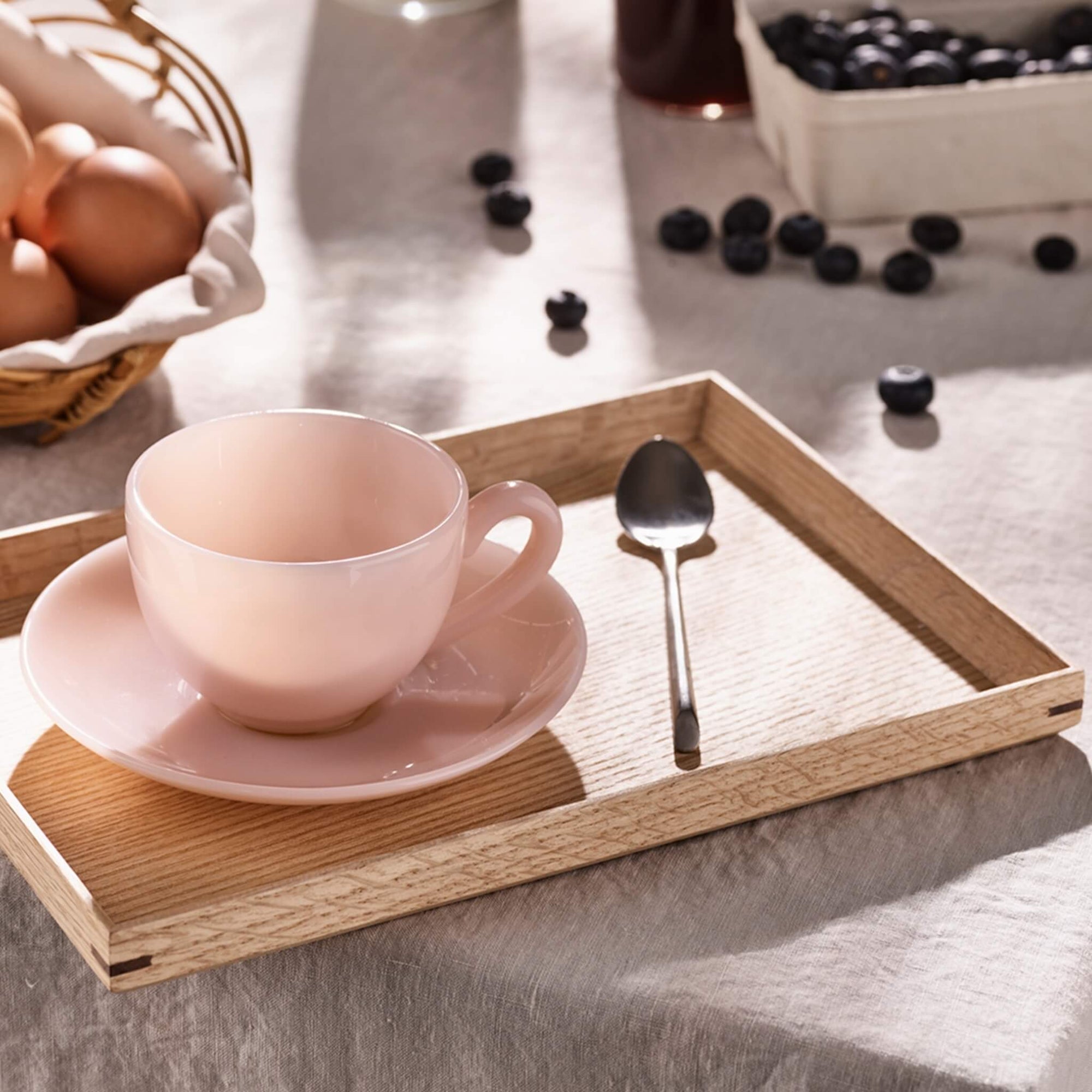 Eine rosa Teetasse mit Untertasse und Löffel auf einem Holztablett, umgeben von Eiern und Blaubeeren.