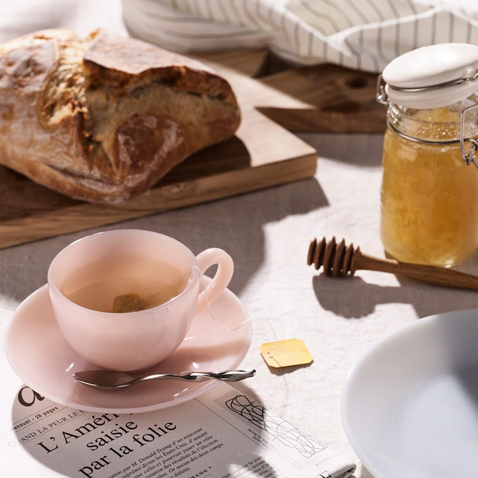 Eine rosa Teetasse mit Tee, ein Löffel, ein Laib Brot, ein Honigglas und eine Zeitung auf einem Tisch.
