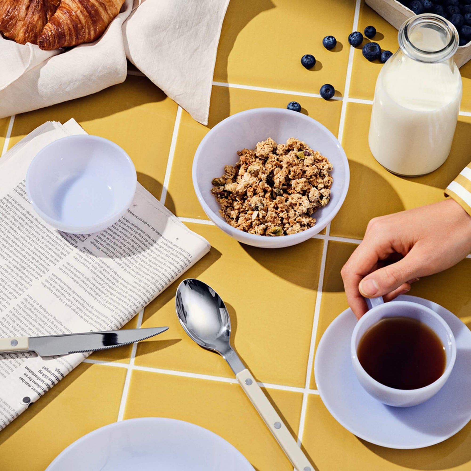 Eine Frühstücksszene mit Müsli, Tee, einem Croissant, Blaubeeren und einer Zeitung auf einem gelb gekachelten Tisch.