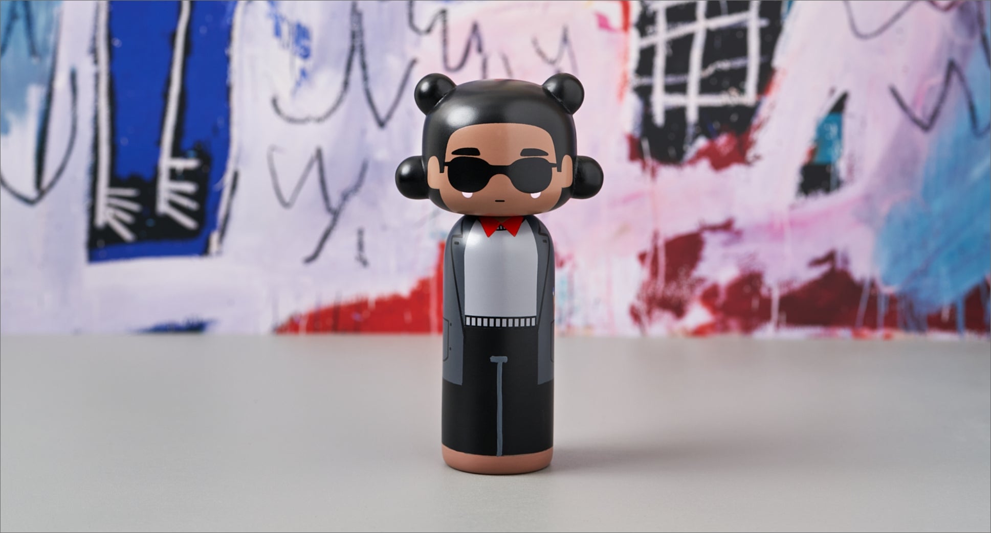 Eine Kokeshi-Puppe mit Sonnenbrille und rotem Hemd vor einem bunten, abstrakten Hintergrund.