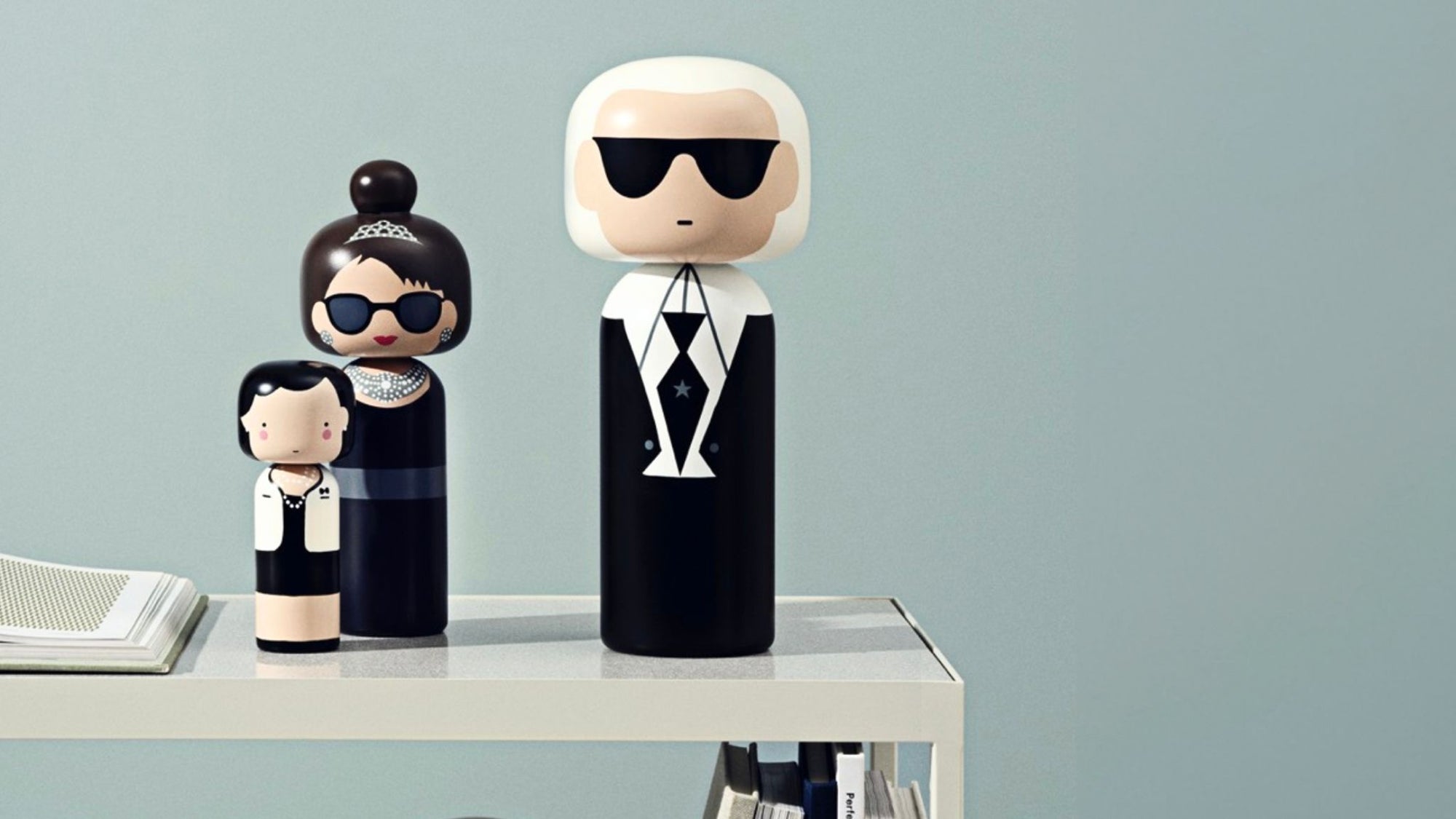 Kokeshi-Puppen in schwarzem und weißem Outfit auf einem Regal vor hellblauem Hintergrund.