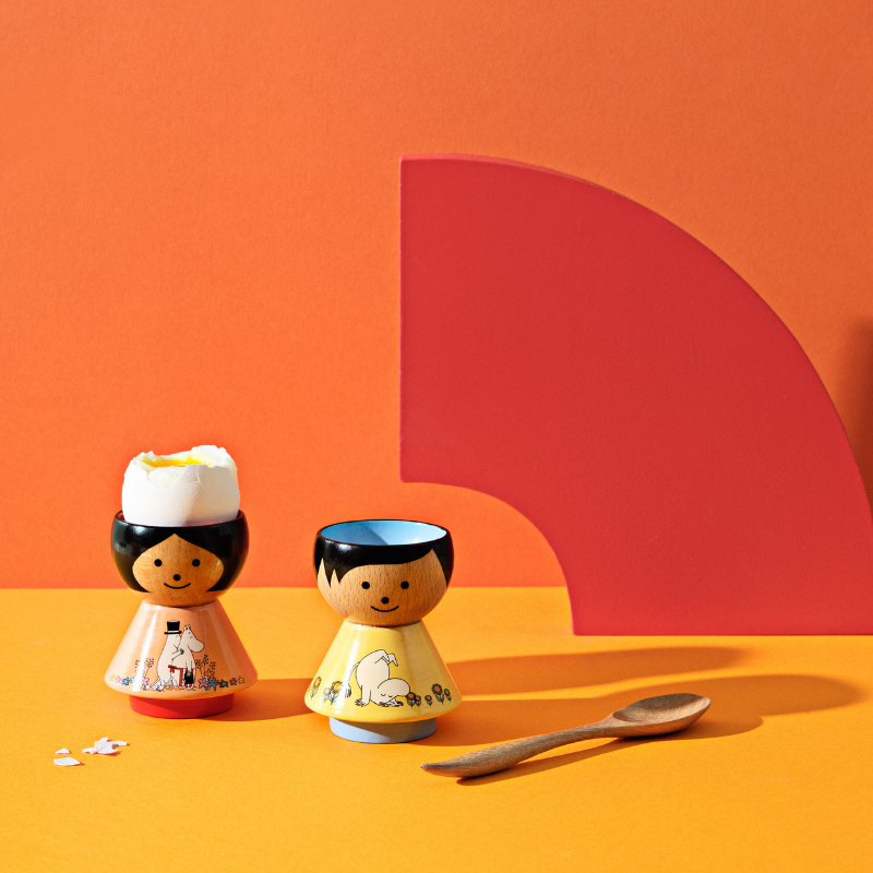Zwei Kokeshi-Puppen-Eierbecher mit einem Holzlöffel auf einer orangefarbenen Oberfläche und rotem Hintergrund.