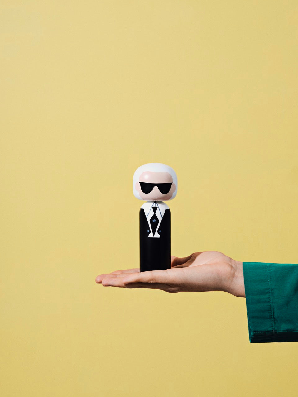 Eine Hand, die eine Kokeshi-Puppe mit Sonnenbrille und einem Anzug vor einem gelben Hintergrund hält.