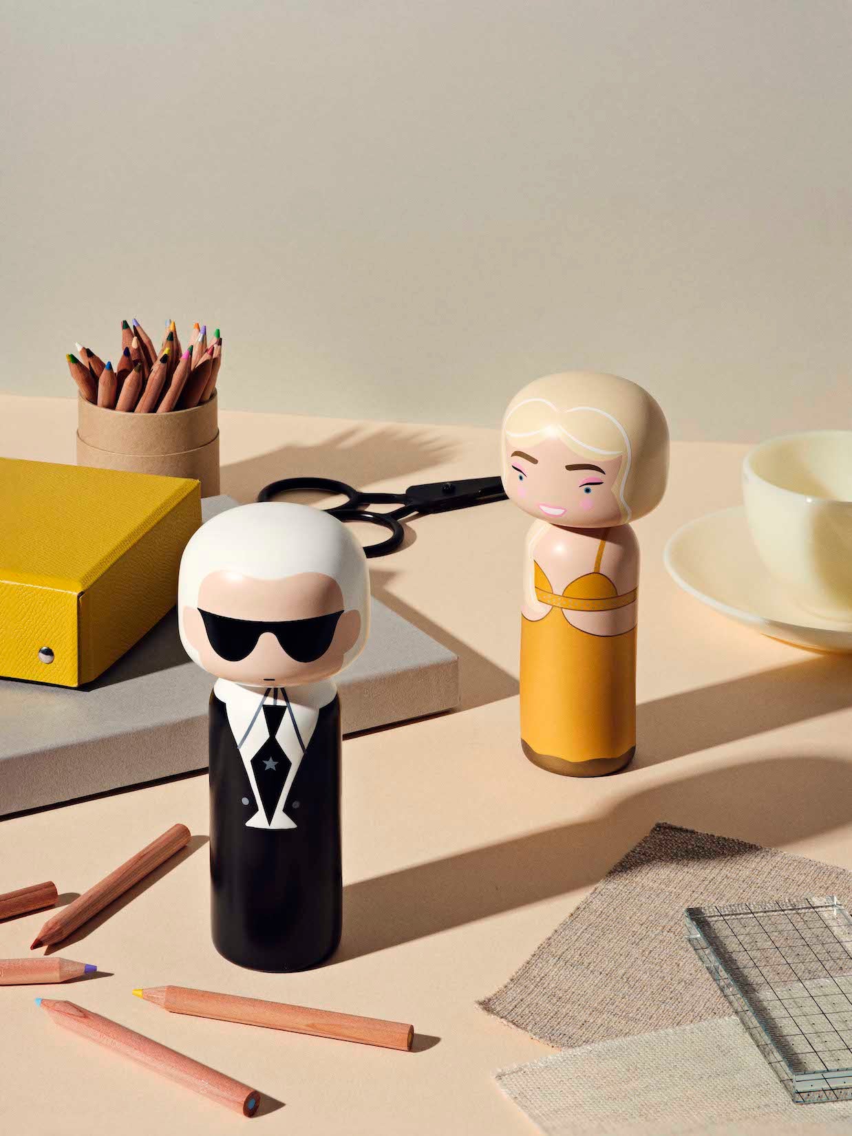 Zwei Kokeshi-Puppen auf einem Tisch mit Buntstiften, einem gelben Hefter und einer Teetasse.