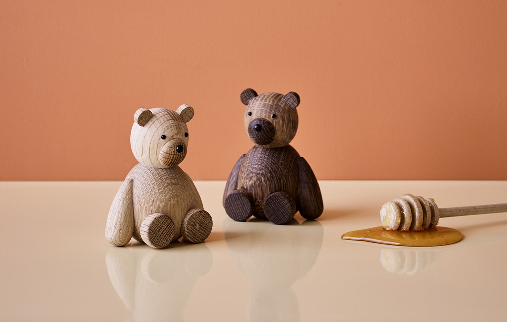 Zwei hölzerne Bärenfiguren sitzen auf einem Tisch neben einem Honiglöffel mit Honig vor einem orangefarbenen Hintergrund.
