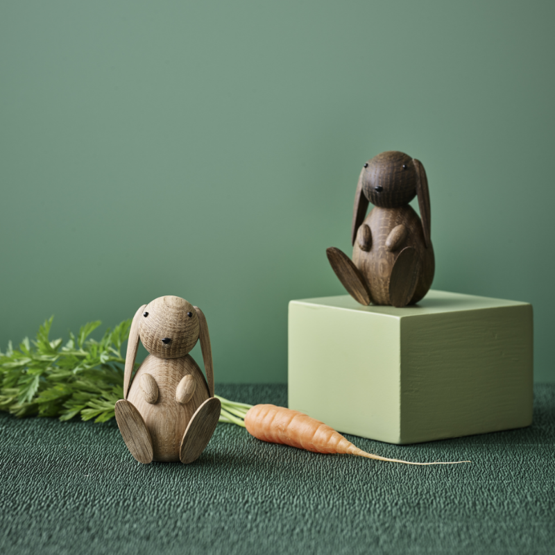 Zwei Kaninchenfiguren aus Holz, eine auf einem grünen Block, mit einer Karotte und Grünzeug auf einer strukturierten Oberfläche.