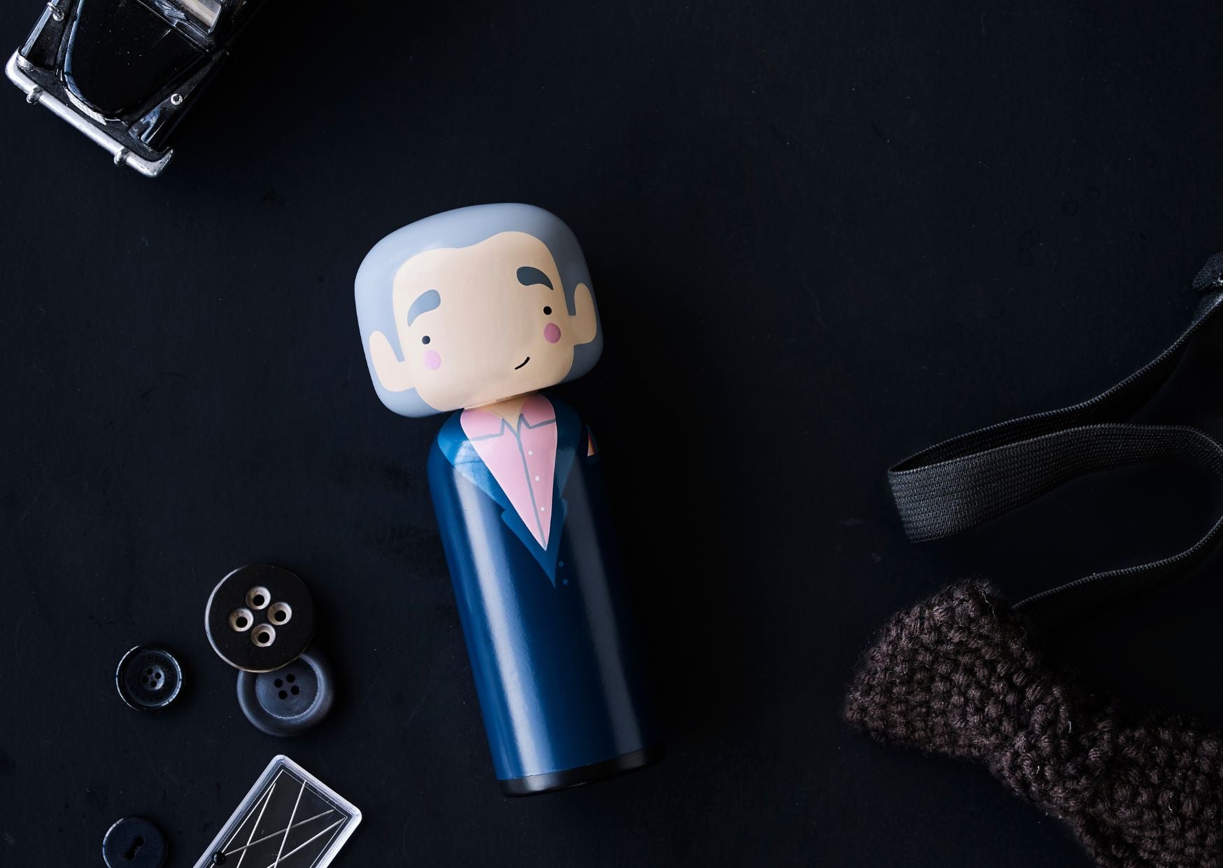 Eine Kokeshi-Puppe mit blauem Anzug und rosafarbenem Hemd auf einer dunklen Fläche, umgeben von Knöpfen und einem gestrickten Gegenstand.