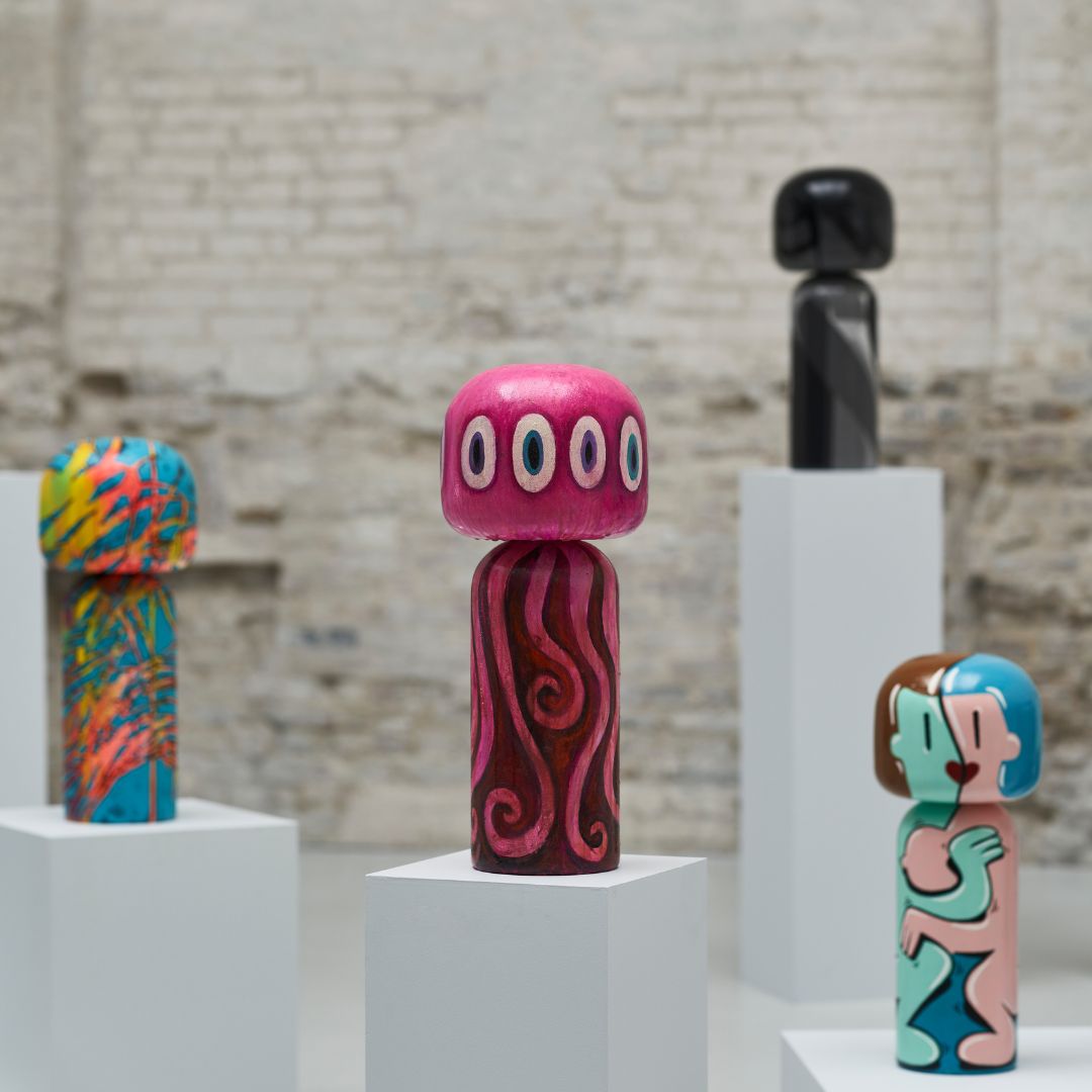 Bunte Kokeshi-Puppen auf weißen Sockeln vor einem Backsteinmauer-Hintergrund.