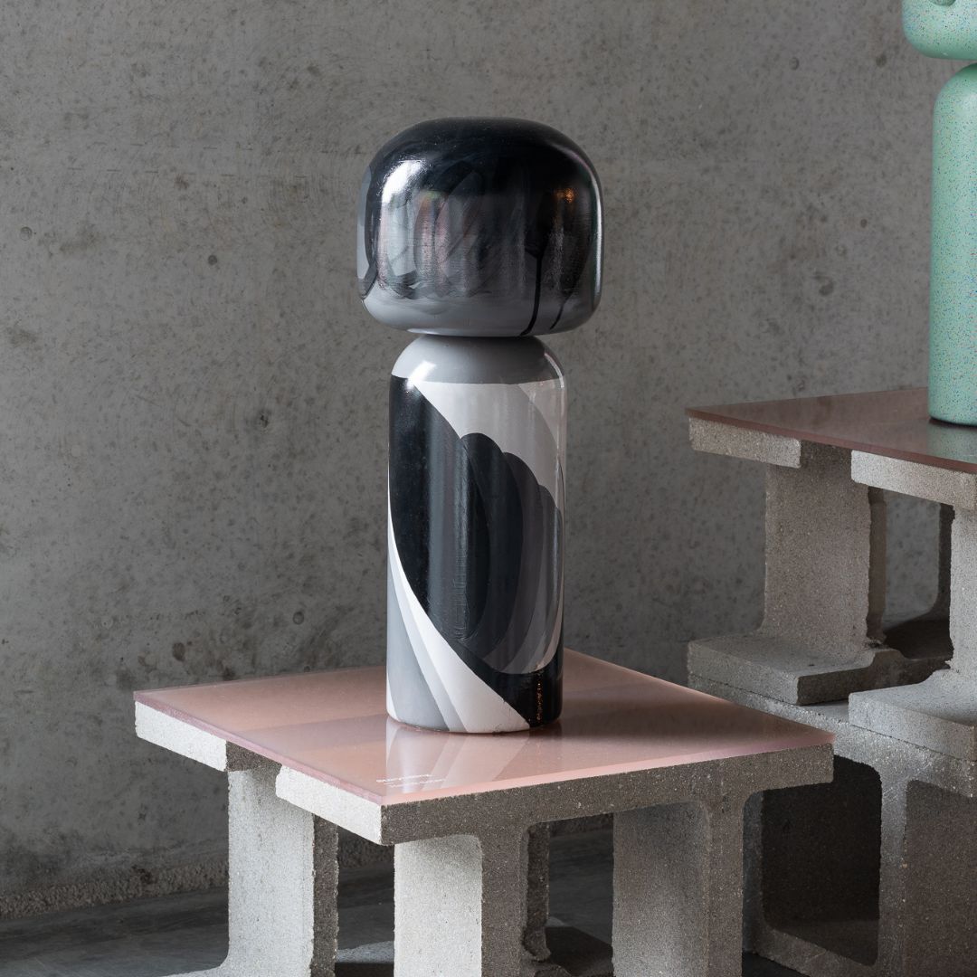 Eine schwarz-graue Kokeshi-Puppe auf einem rosa Tisch aus Betonblöcken vor einer grauen Wand.