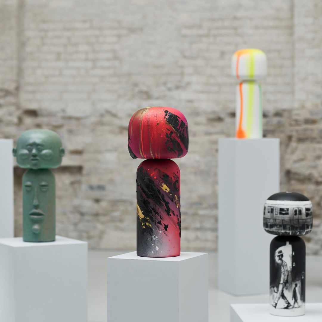 Bunte Kokeshi-Puppen auf weißen Sockeln vor einem Backsteinmauer-Hintergrund.