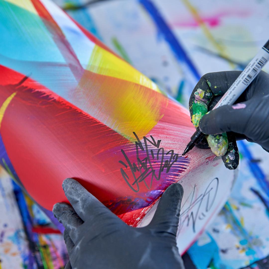Künstler mit Handschuhen signiert ein buntes, abstrakt gemaltes Objekt auf einer bespritzten Oberfläche.