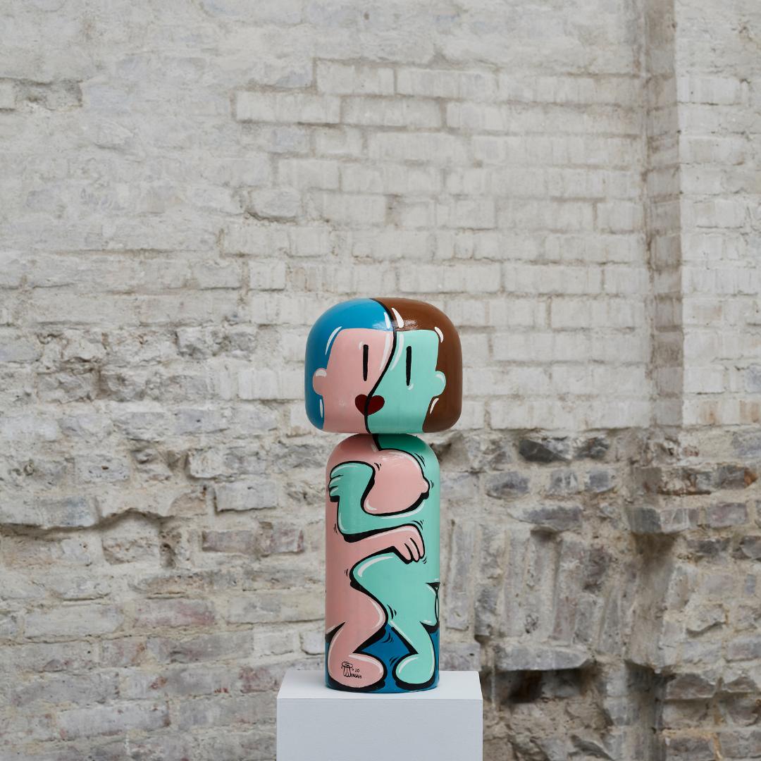 Eine farbenfrohe Kokeshi-Puppe mit abstraktem Muster auf einem weißen Sockel vor einer strukturierten Ziegelwand.