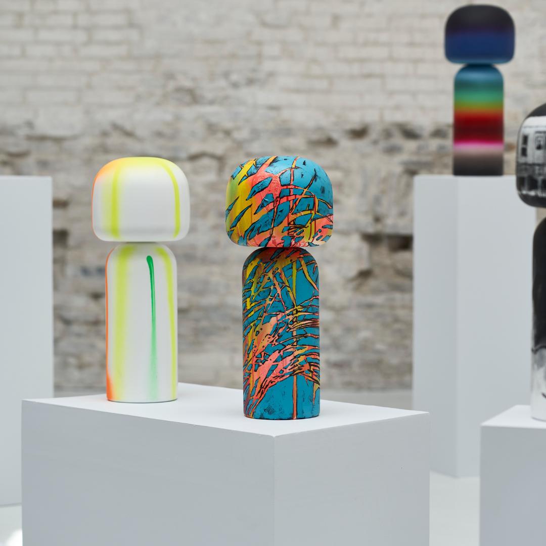 Bunte Kokeshi-Puppen auf weißen Sockeln vor einem strukturierten Backsteinmauer-Hintergrund.