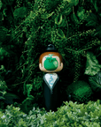Une poupée kokeshi avec un visage en forme de pomme verte se tient au milieu d'un feuillage vert luxuriant.