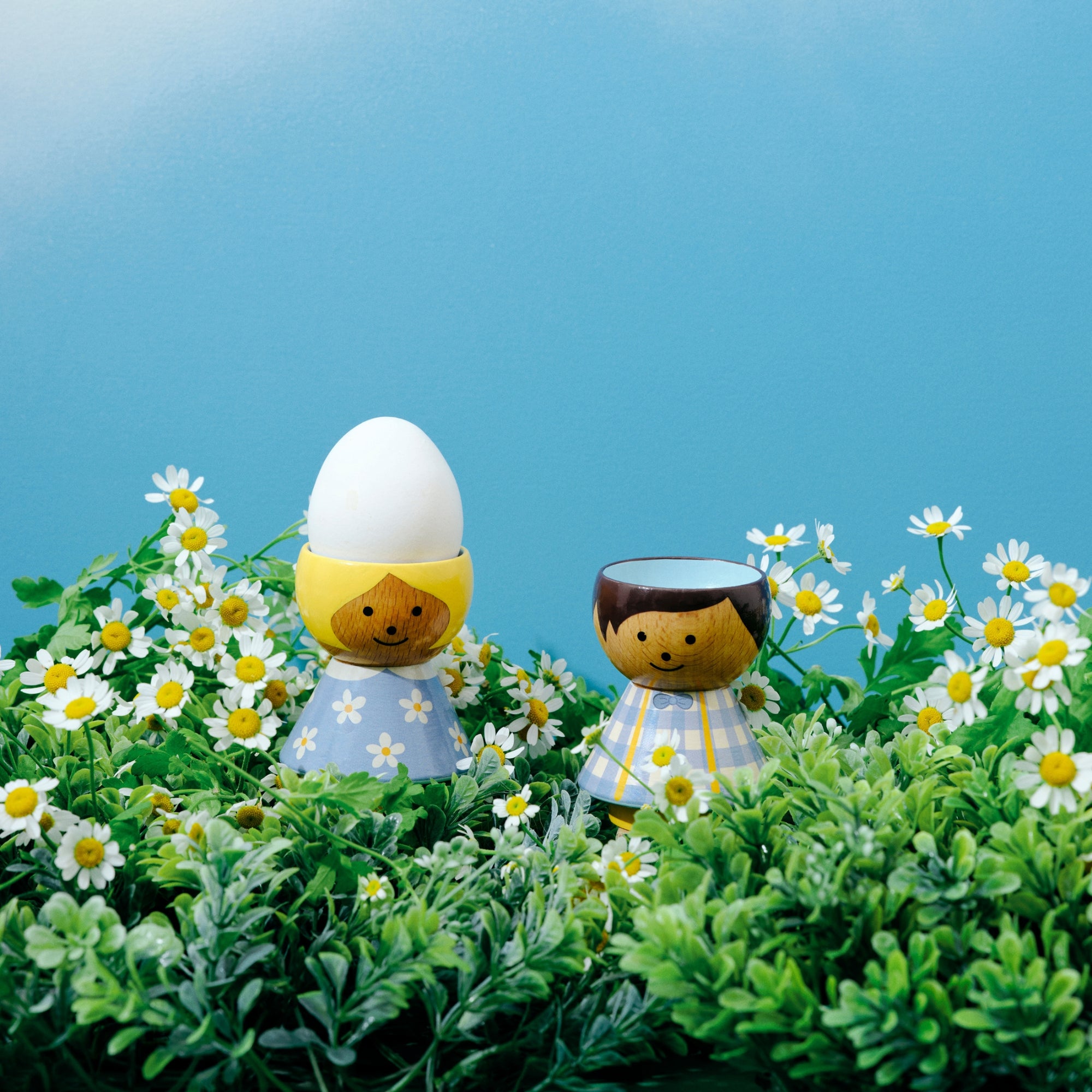 Poupées Kokeshi dans un champ de marguerites, dont une avec un œuf sur la tête, sur fond de ciel bleu.