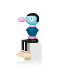 Figure abstraite colorée en bois avec un visage bleu, un nez rose et un corps géométrique assis sur un bloc blanc.