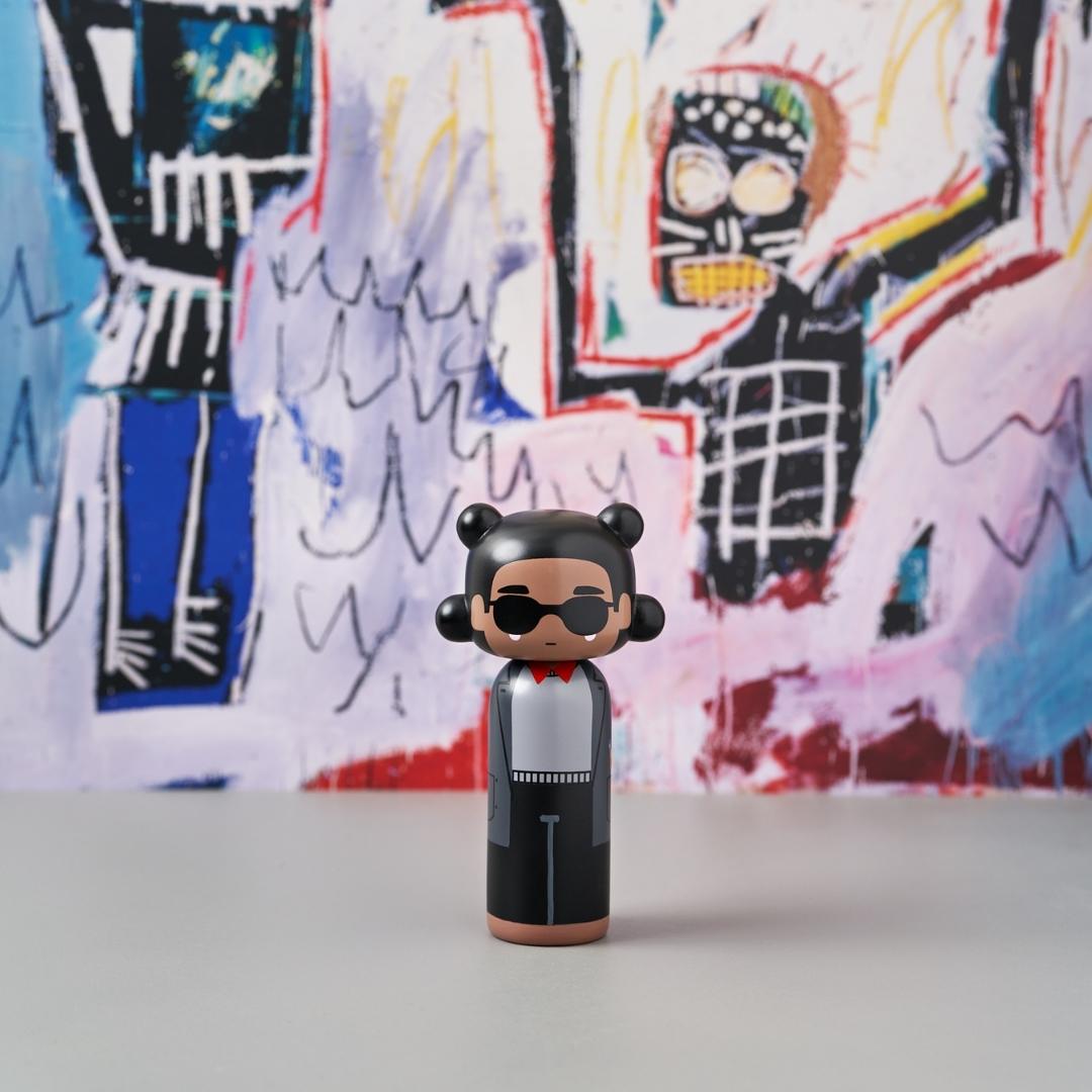 Poupée Kokeshi avec lunettes de soleil et tenue noire devant une peinture abstraite colorée.