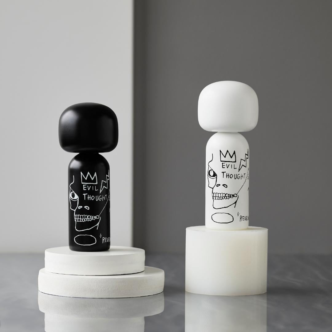 Deux poupées kokeshi, l'une noire et l'autre blanche, avec des motifs artistiques, sur des piédestaux ronds sur fond gris.