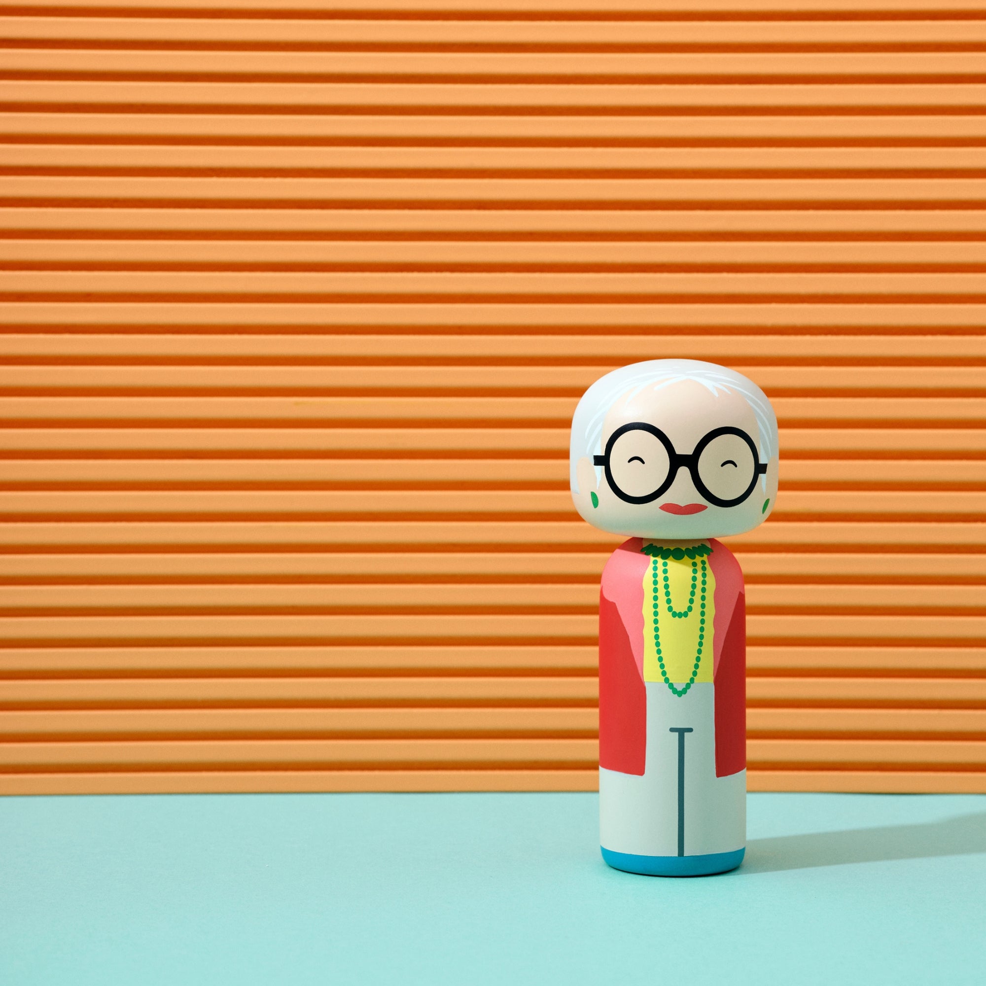 Une poupée kokeshi avec des lunettes et une veste rouge sur un fond rayé orange.